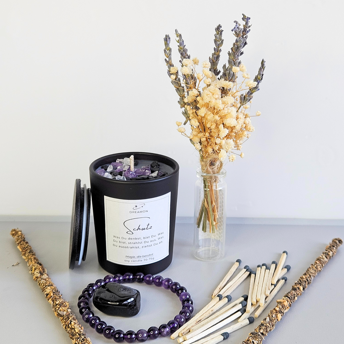 Detailaufnahme Schutzset: schwarze Ritualkerze, Glas mit Streichhölzern, Trockenblumenstrauß, Amethyst-Armband, Turmalin und Räucherstäbchen.