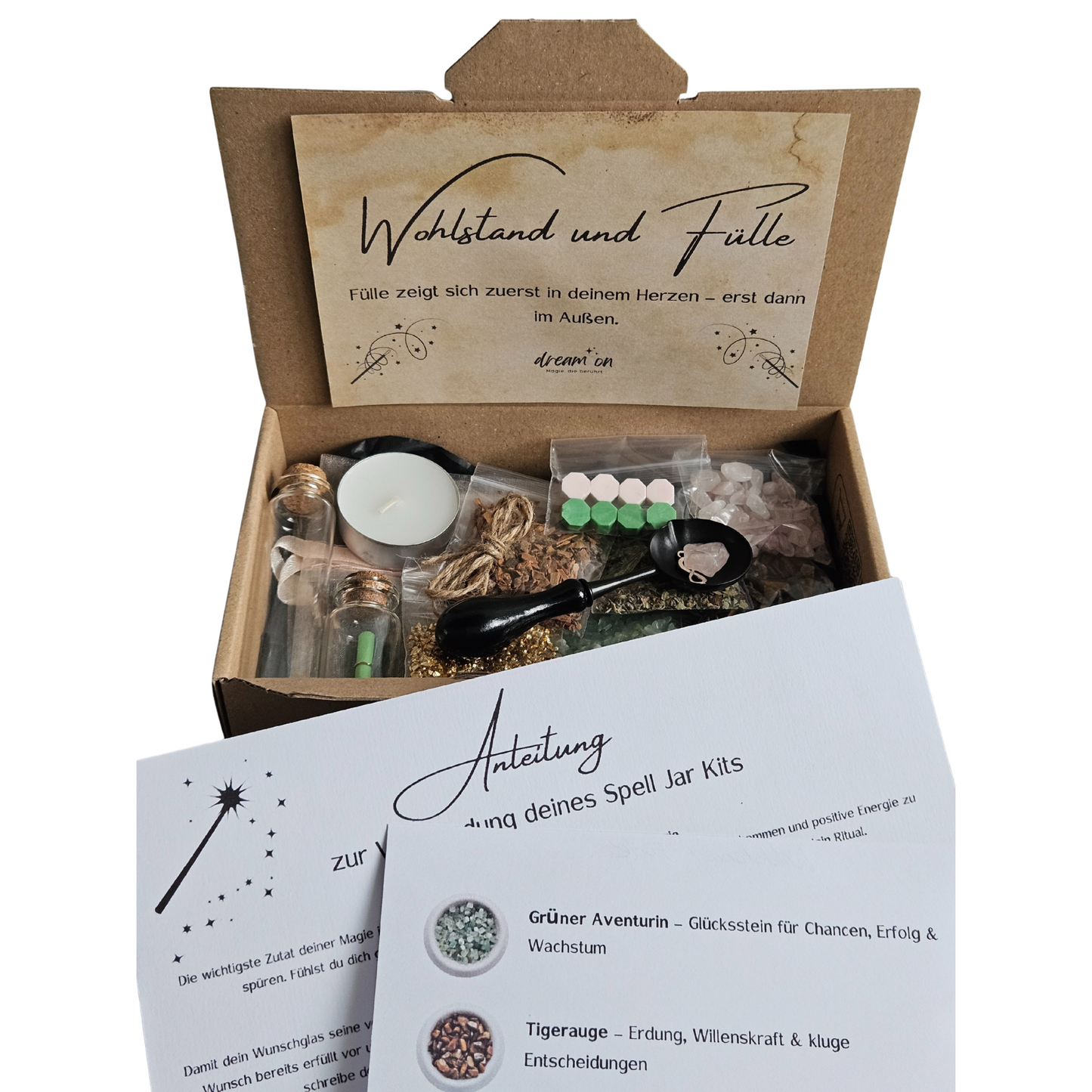 Geöffnete Geschenkbox des DIY-Kreativsets Fülle von DreamOn, gefüllt mit Naturmaterialien und Zubehör. Im Vordergrund liegt eine gedruckte Anleitung zur Verwendung des Sets mit Beschreibungen zu Grünem Aventurin, Tigerauge und Goldflocken.