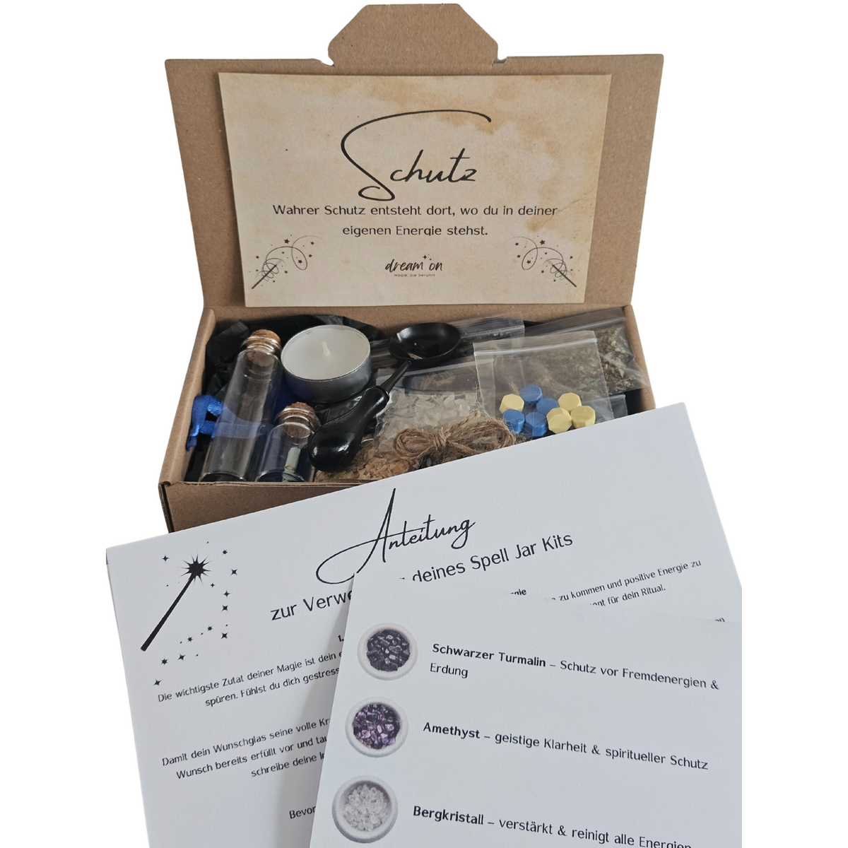 Geöffnete Geschenkbox des DIY-Kreativsets Schutz von DreamOn, gefüllt mit Naturmaterialien und Zubehör. Im Vordergrund liegt eine gedruckte Anleitung zur Verwendung des Sets mit Beschreibungen zu Schwarzem Turmalin, Amethyst und Bergkristall.