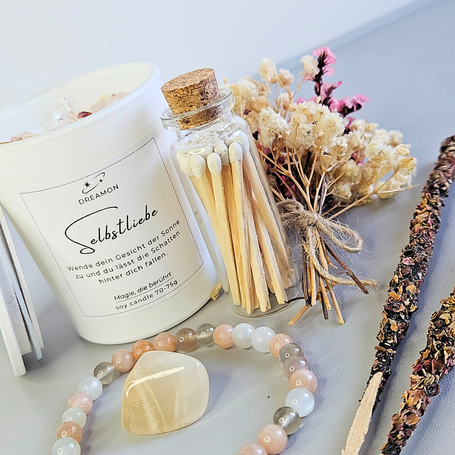 Detailaufnahme Ritualset Selbstliebe: weiße Duftkerze, Glasröhrchen mit Streichhölzern, Trockenblumen, Mondstein-Armband, Edelstein und Räucherstäbchen.