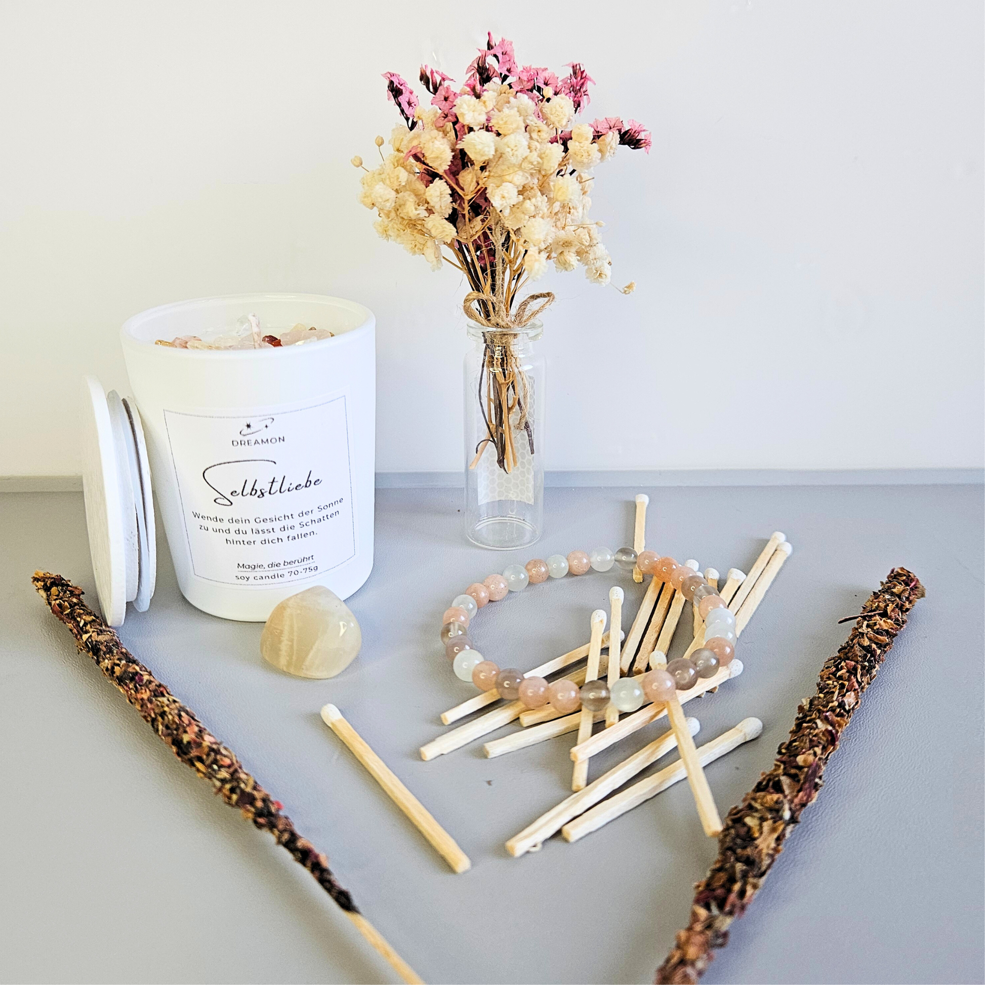 Geschenkset Selbstliebe arrangiert: Kerze mit Heilsteinen, Mondstein-Armband, Edelstein, Trockenblumen, Streichhölzer und zwei Rosen-Olibanum Räucherstäbchen.