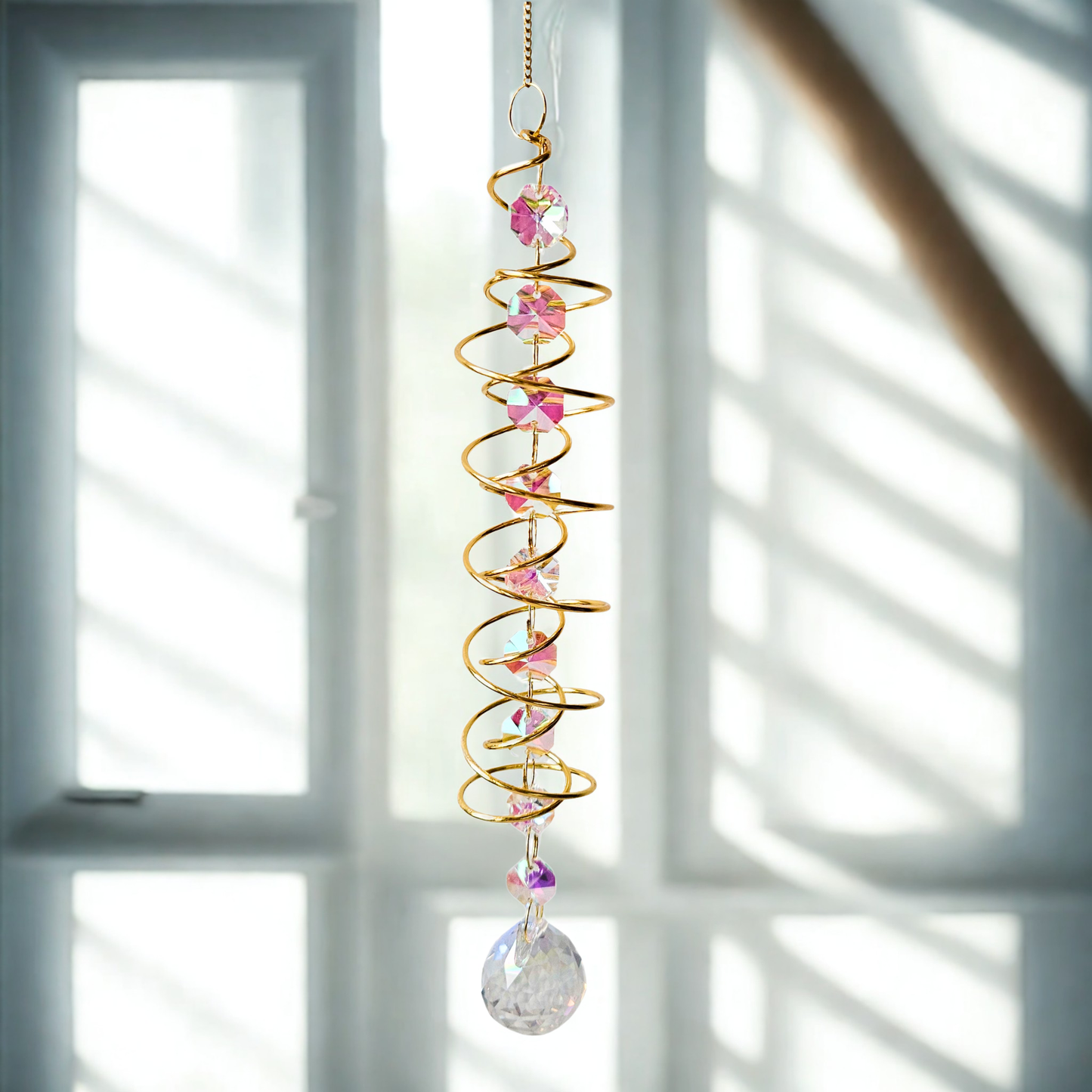 Goldener Spiral-Suncatcher mit irisierenden Kristallperlen und großem Prisma, dekorativ hängend vor einem Fenster im sanften Tageslicht.