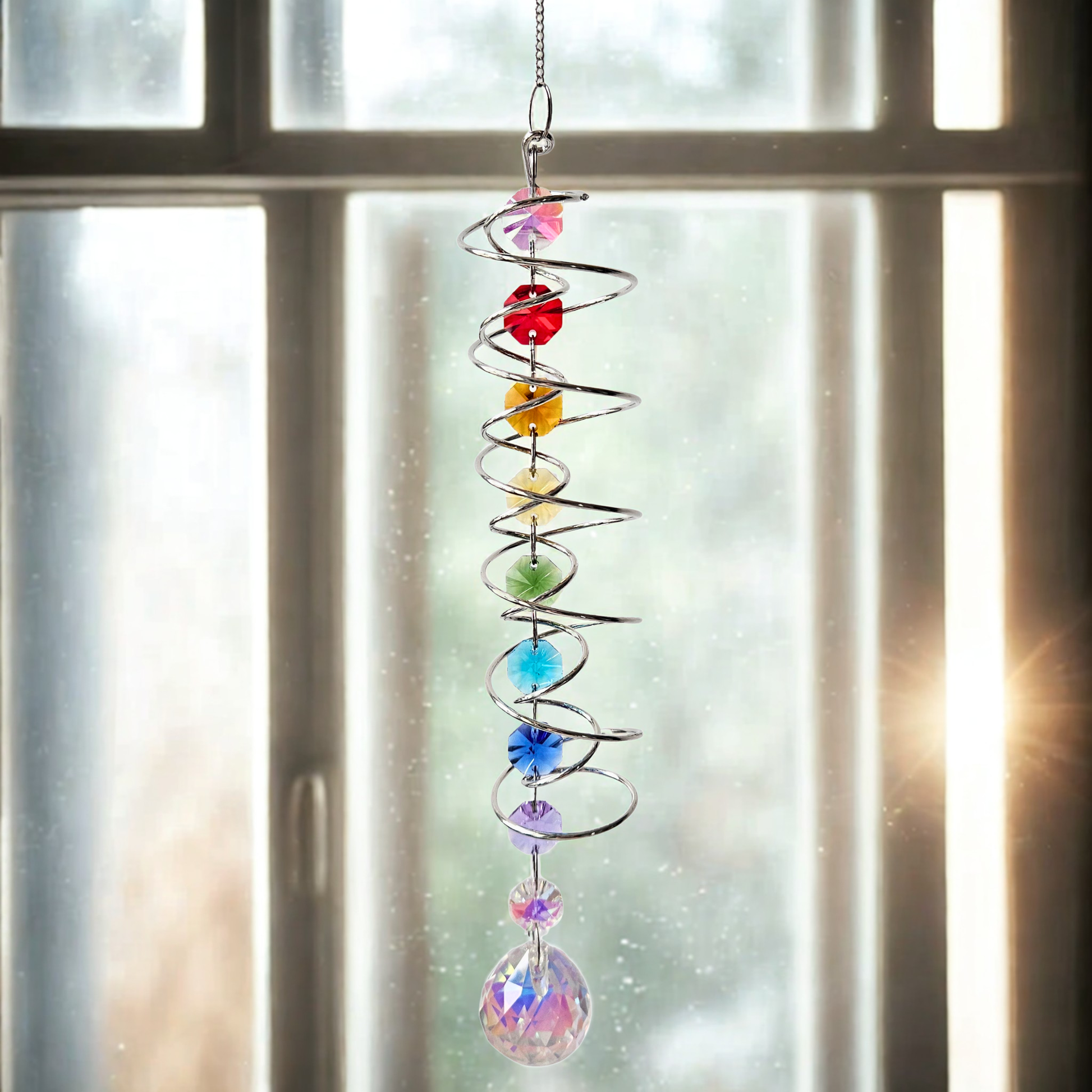 Silberner Chakra-Suncatcher mit farbigen Kristallperlen und grossem Regenbogenprisma, hängend vor einem Fenster im weichen Tageslicht.