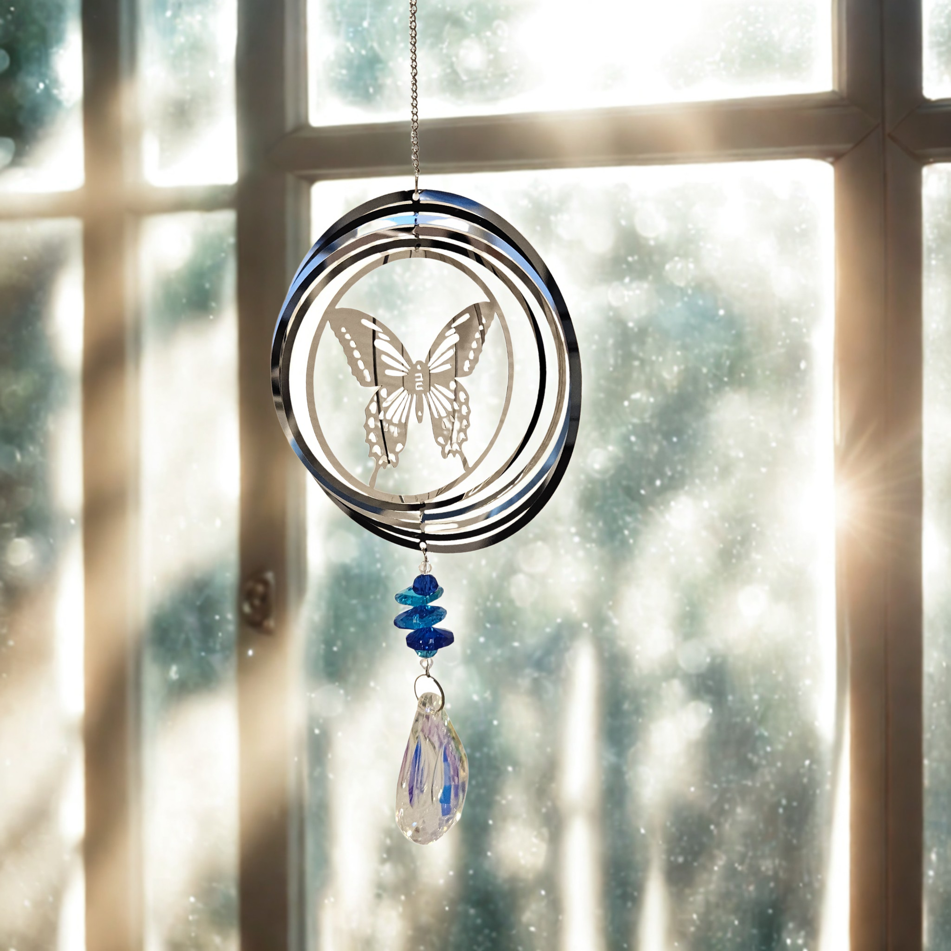 Metall-Suncatcher Windspiel mit Schmetterling und blauem Kristalltropfen, hängend im Sonnenlicht vor einem Fenster.