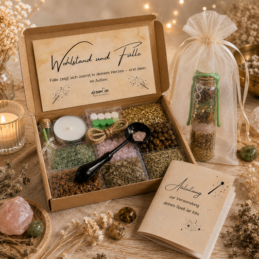 Geöffnete Geschenkbox des DIY-Kreativsets Wohlstand und Fülle von DreamOn mit Naturmaterialien, Kristallen, Goldflocken und Zubehör zum Versiegeln. Rechts steht ein fertig gestaltetes Wunschglas mit grünem Wachssiegel im hellen Organzasäckchen, im Vordergrund liegt das Anleitungsheft.
